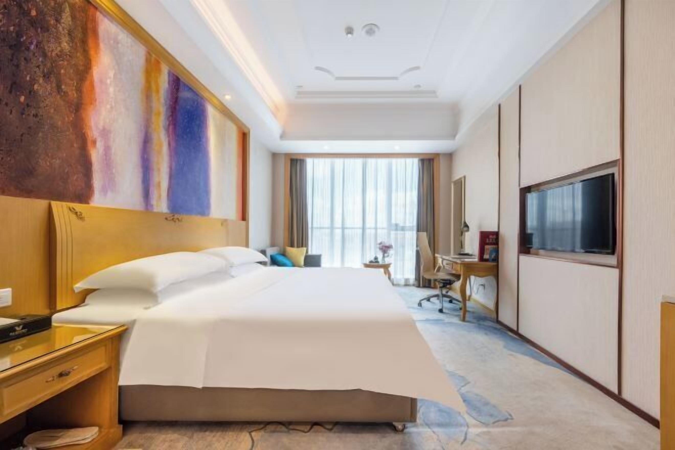 A hotel room with a large bed, a desk with a chair, a television, and a large window with curtains.