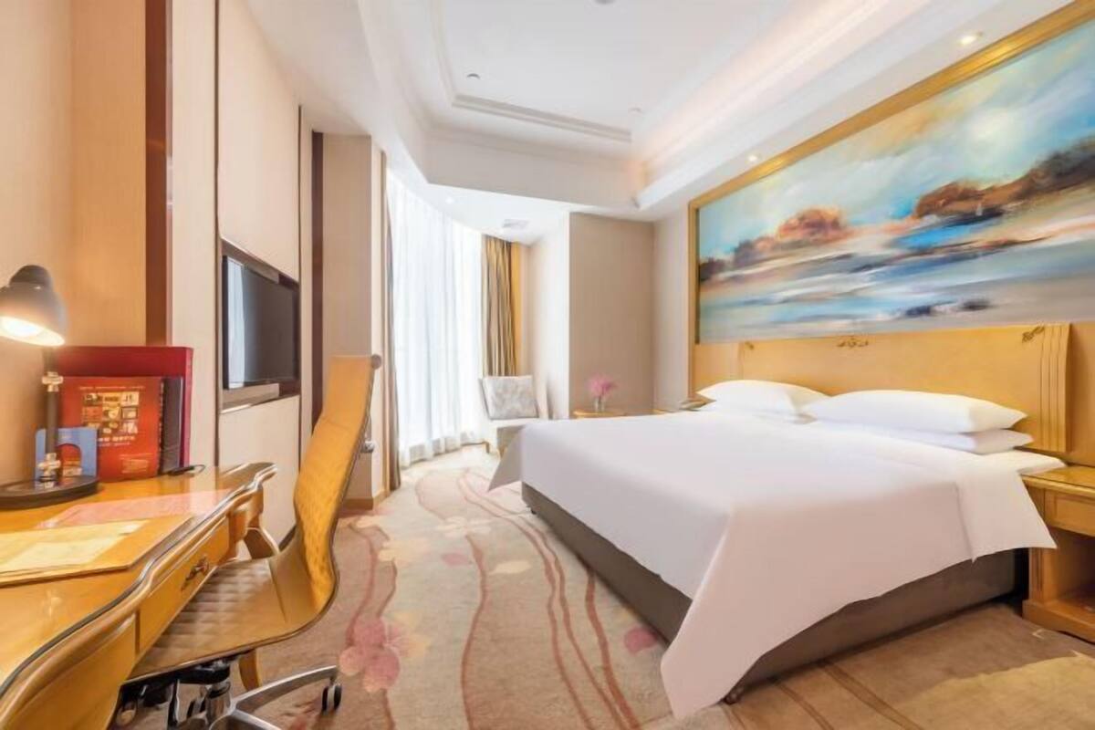 A hotel room with a bed, desk, chair, and a painting on the wall.