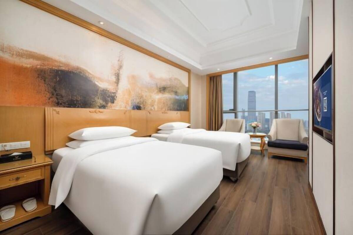 A hotel room with two beds, a large abstract painting on the wall, a view of the city through large windows, and a small table with a vase of flowers.