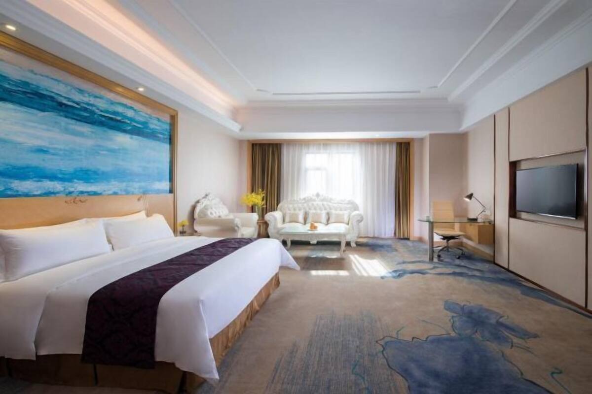 A hotel room with a large bed, a desk, a chair, a TV, and a painting on the wall.