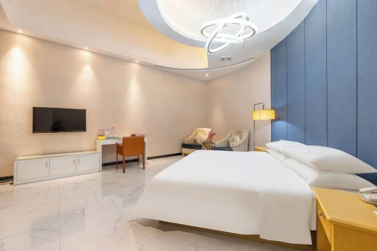 A modern hotel room with a large bed, a desk, a chair, a TV, and a bedside table with a lamp.