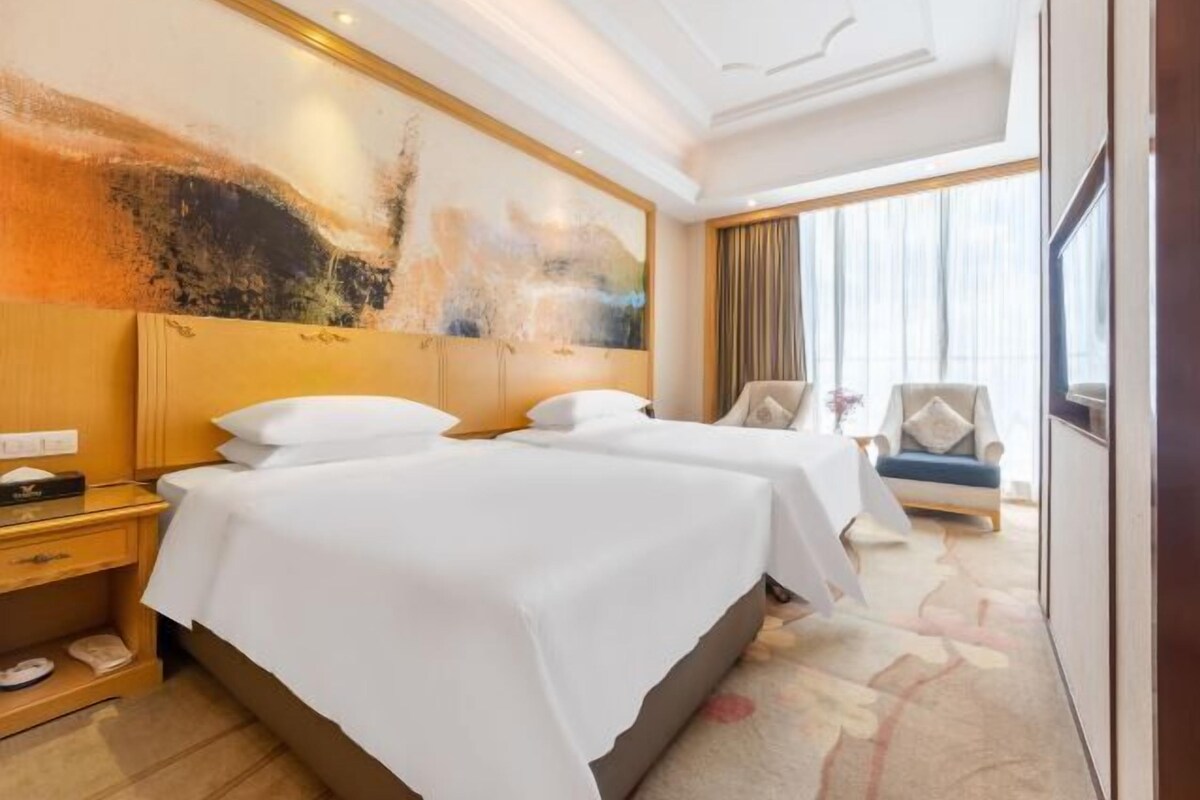 A hotel room with two beds, a large abstract painting on the wall, a chair, and a view of the city through the window.