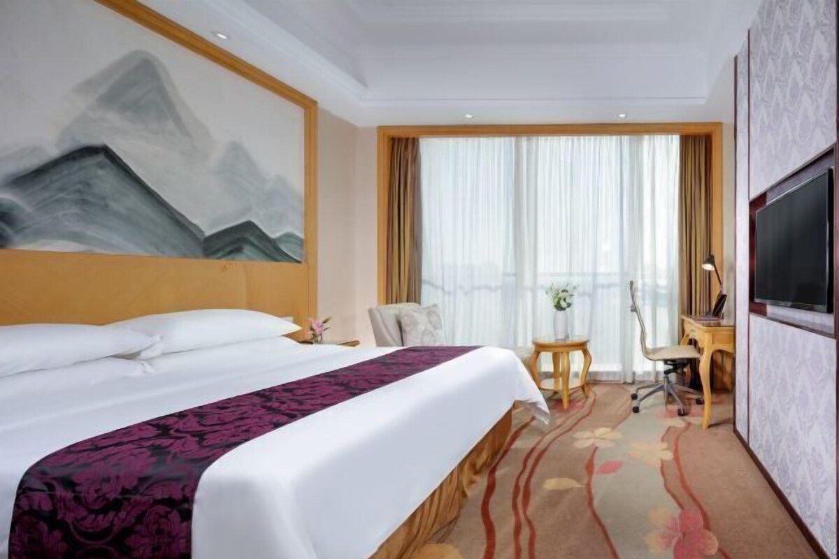A hotel room with a large bed, a desk, a chair, a TV, and a painting on the wall.