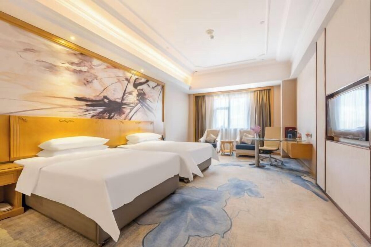 A hotel room with a large bed, a desk, a chair, and a large window with curtains.