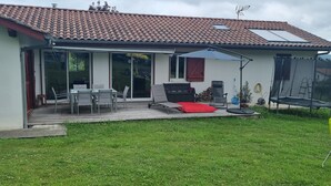 A house with a tiled roof, a patio with a table and chairs, a red cushion, a trampoline, and a covered area with a chair and umbrella. - Maison Chaleureuse Avec Piscine Entre Montagne ET MER (ASCAIN)