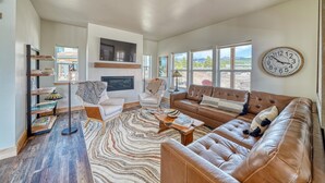 A spacious living room with a large brown leather sofa, a white armchair, and a coffee table. A large wall clock and a fireplace are also visible. - Silver Rush- Brand New Railyard home minutes to Copper & Ski Cooper! (Leadville)