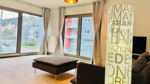 Ein modernes Wohnzimmer mit großem Fenster, einer Couch, einem Hocker und einem an der Wand montierten Fernseher.