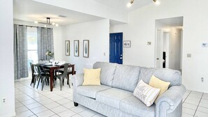 Ein modernes Wohnzimmer mit einem grauen Sofa, einem Esstisch und Stühlen. Eine blaue Tür und ein gerahmtes Bild sind an der Wand zu sehen.