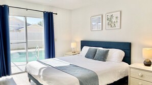 Ein Schlafzimmer mit Bett, Nachttisch, Lampe, Fenster mit Vorhängen und Blick auf einen Swimmingpool.