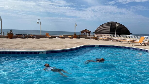 Una piscina amb dues persones nedant, una zona de platja amb gandules i una gran estructura de cúpula al fons.
