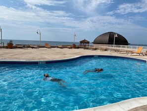 A swimming pool with two people swimming, a beach area with lounge chairs, and a large dome structure in the background.