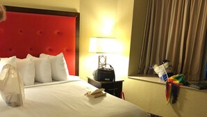 Habitació d'hotel amb un panell de capçal de llit vermell, un llit amb roba de llit blanca, una làmpada de nit, una taula de nit amb una làmpada i una finestra amb cortines, tot sobre un sòl amb moqueta.