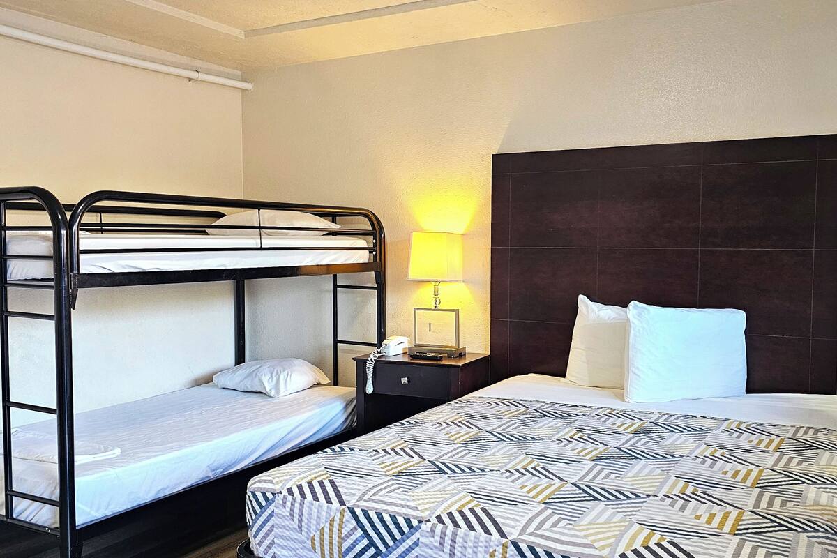 A hotel room with bunk beds, a bedside table with a lamp, and a large bed with a patterned bedspread.