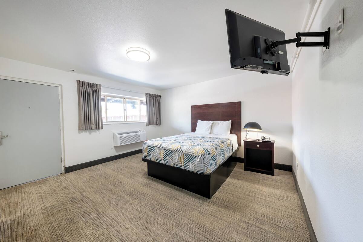 A hotel room with a bed, a TV mounted on the wall, a bedside table, and a window with curtains.