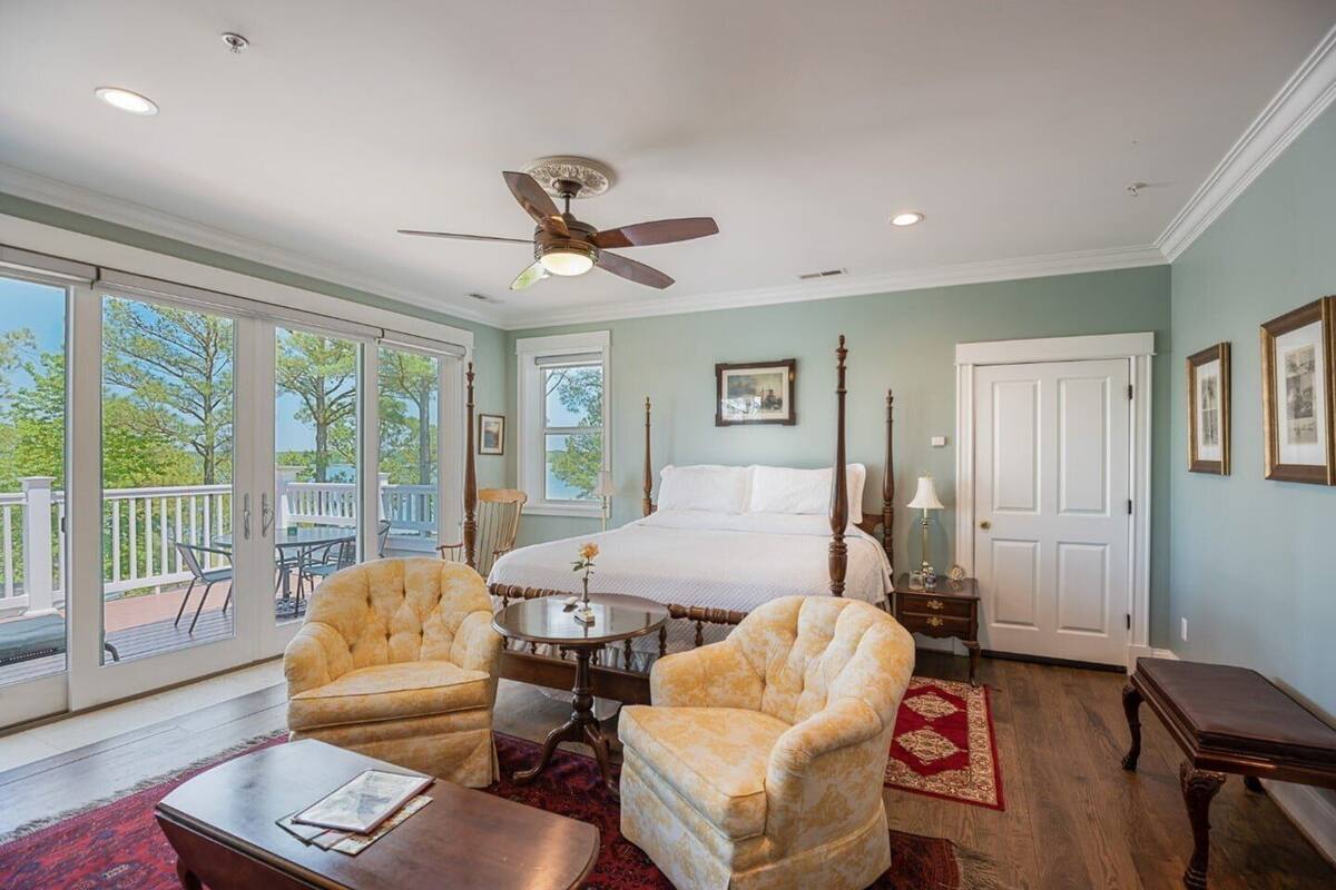 A bedroom with a large bed, two armchairs, a small table, a ceiling fan, and a view of the outdoors.