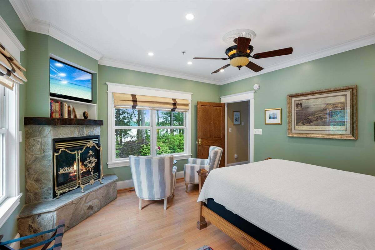 A bedroom with a stone fireplace, a bed, a ceiling fan, and a view of the outdoors.