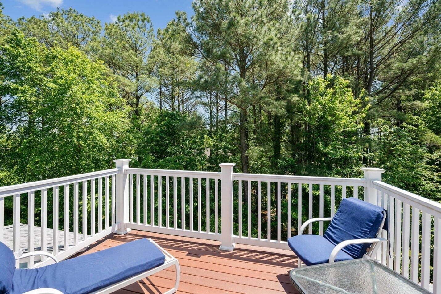 Una terraza de madera con barandas blancas, dos sillones de descanso con cojines azules y una mesa de vidrio.