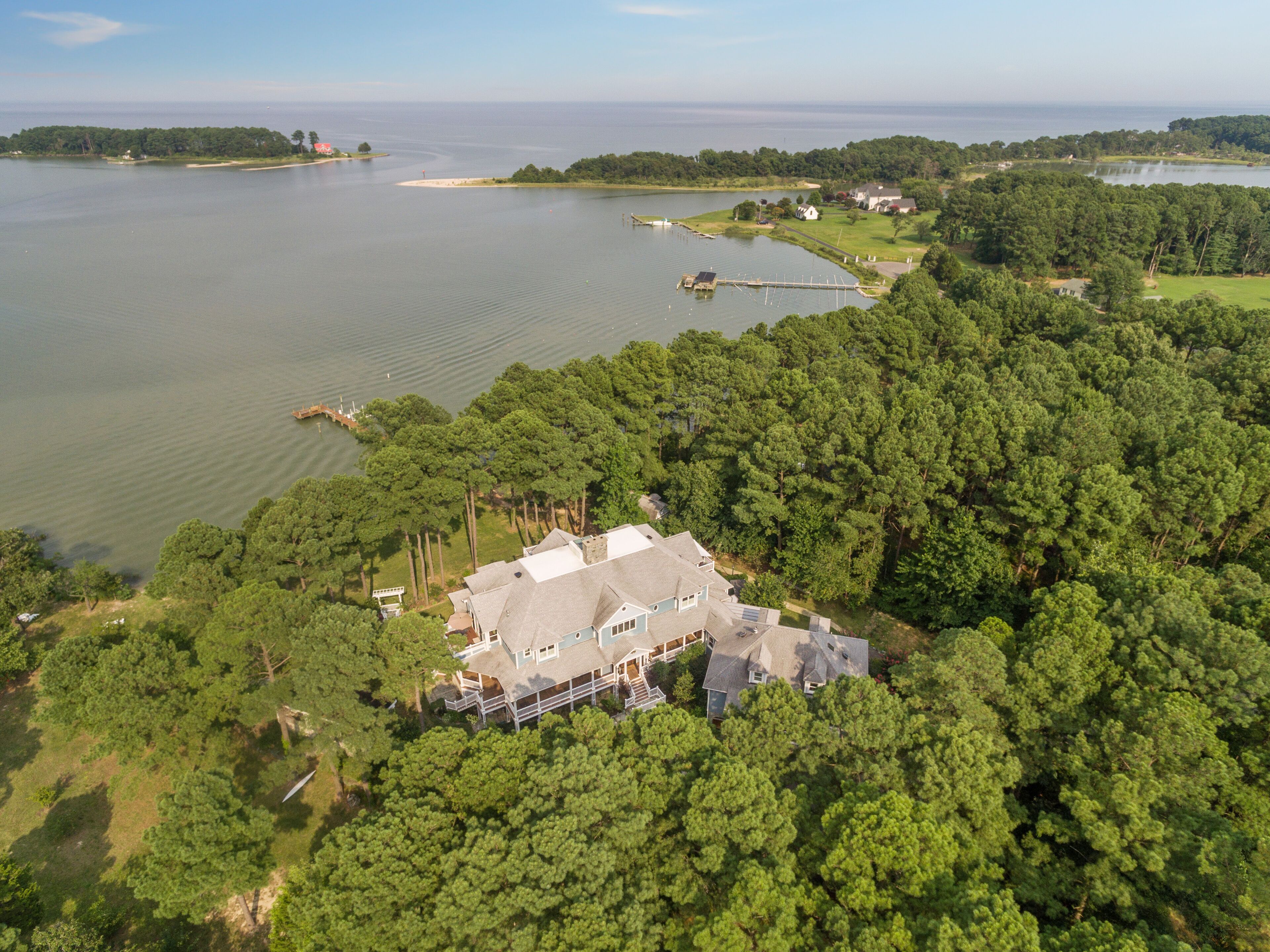 An aerial view of a large house surrounded by dense forest, with a lake and dock in the background.