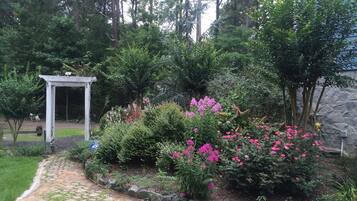 Un jardín con un sendero de piedra, una pérgola blanca y arbustos de flores rosadas.