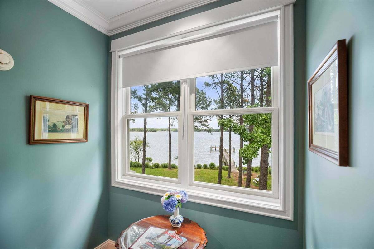 A room with a large window overlooking a lake, framed by a white window sill and a wooden table with a vase of flowers.
