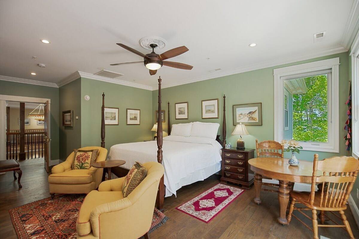 A bedroom with a four-poster bed, a ceiling fan, a dining table with chairs, and a view of greenery through a window.