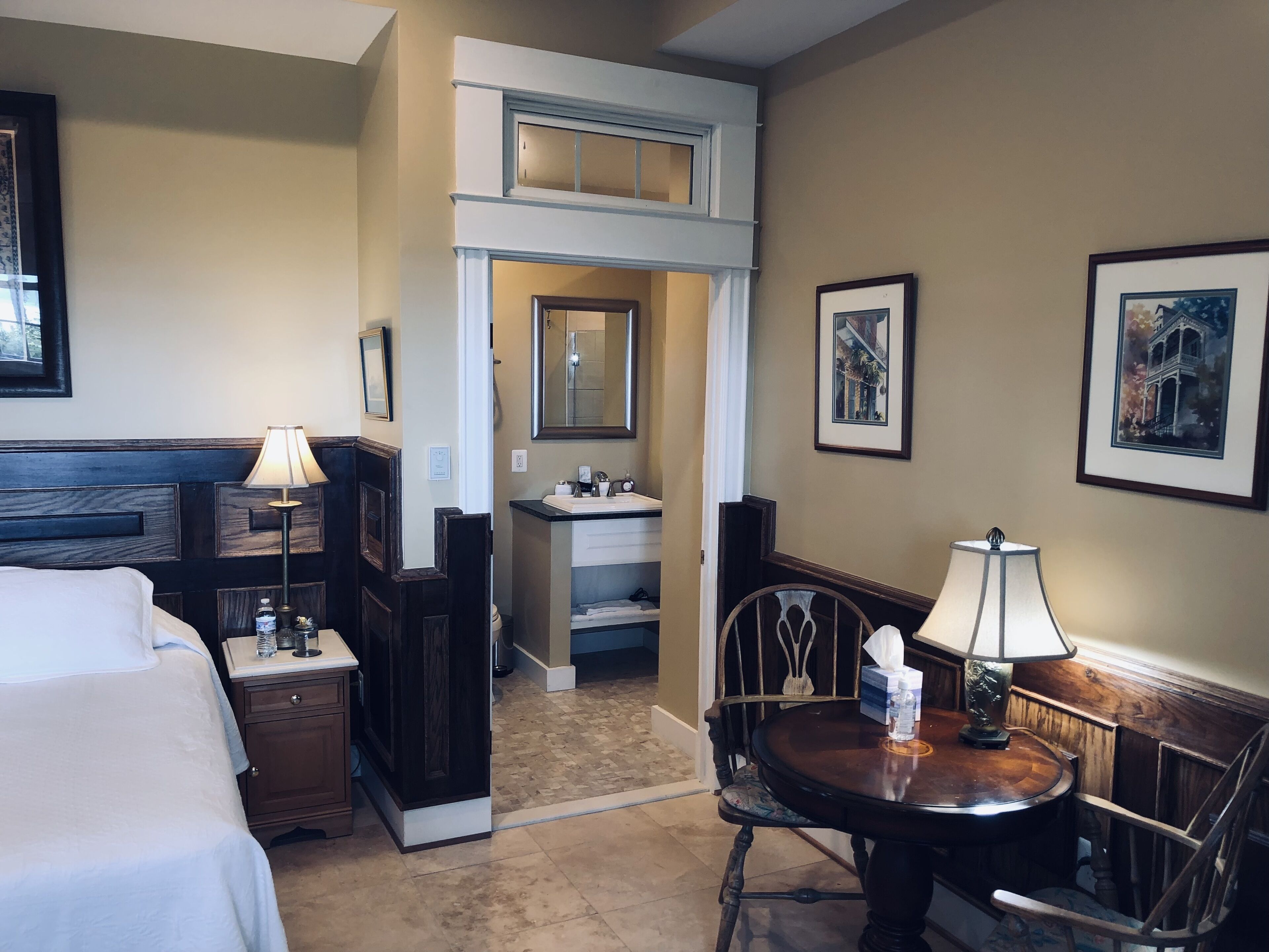 A hotel room with a bed, a desk with a lamp, a chair, a small table with a lamp, a bathroom with a sink, and framed pictures on the wall.