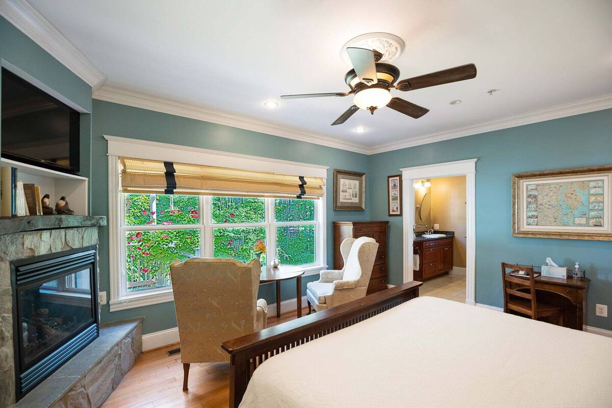 A bedroom with a fireplace, a bed, a desk, and a window with a view of greenery.