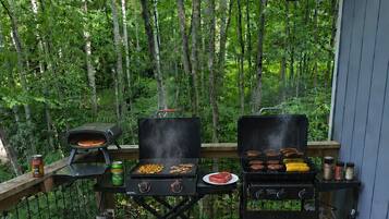 Una terraza de madera con dos parrillas para barbacoa, un bidón de gas y una pequeña mesa.