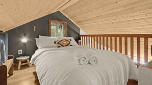 Un dormitorio acogedor con techo de madera, una cama con sábanas blancas, un taburete pequeño, una mesita de noche con lámpara y una ventana con vistas a la vegetación.