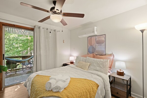 Un dormitorio con cama, ventilador de techo, mesita de noche con lámpara, una hamaca afuera de la ventana y vista a vegetación.