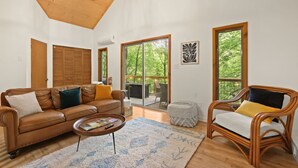 Un salón espacioso con un sofá de cuero marrón, una mesa de centro de madera y un sillón de mimbre. Grandes ventanales ofrecen vistas a la vegetación exterior.