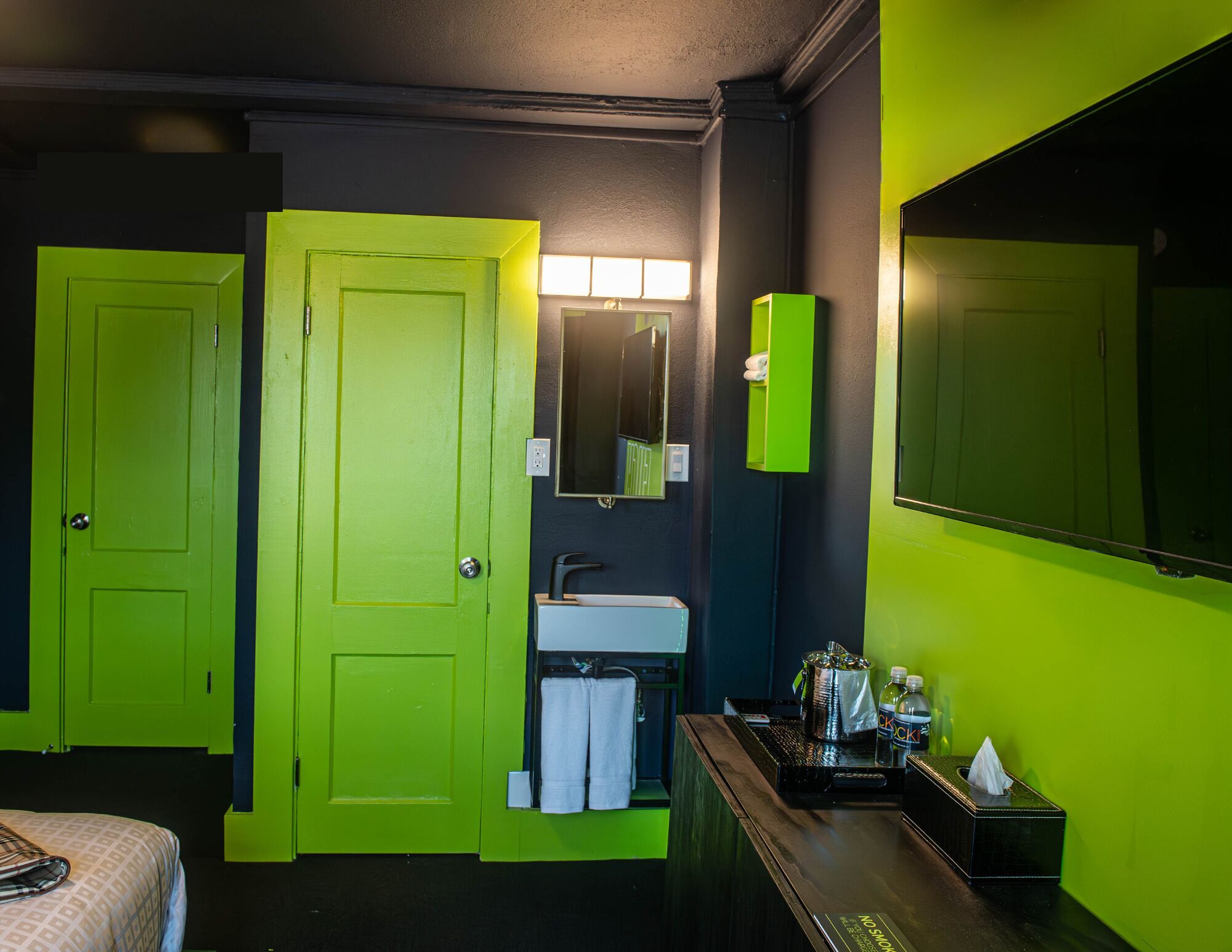 A hotel room with a bright green door, a sink with a mirror above it, a large mirror on the wall, and a bedside table with a tissue box and bottles.