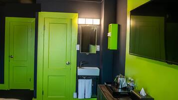 A hotel room with a bright green door, a sink with a mirror above it, a large mirror on the wall, and a bedside table with a tissue box and bottles.