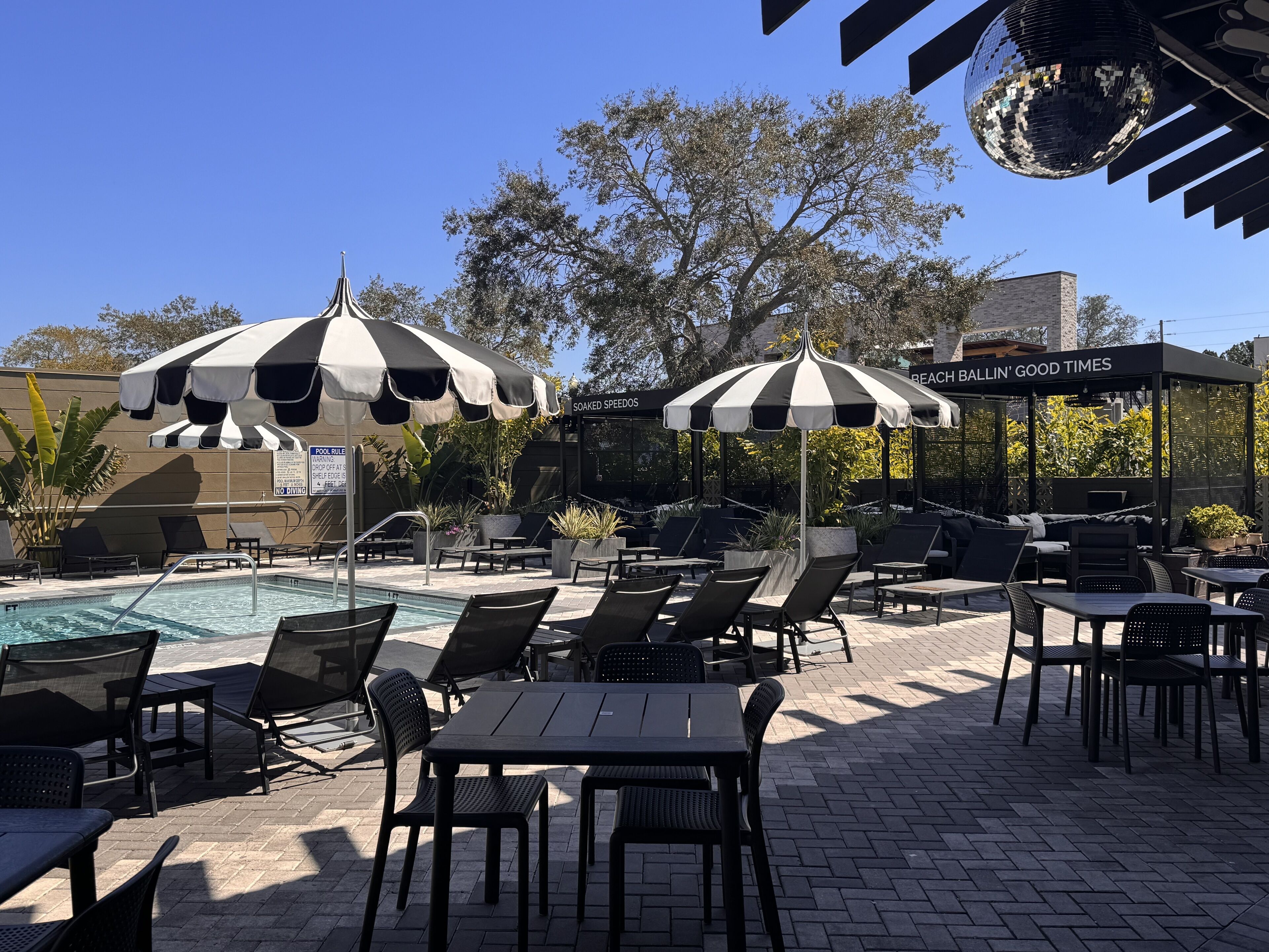 A poolside area with lounge chairs and tables, umbrellas, and a sign for 'Beach Ballin' Good Times'.
