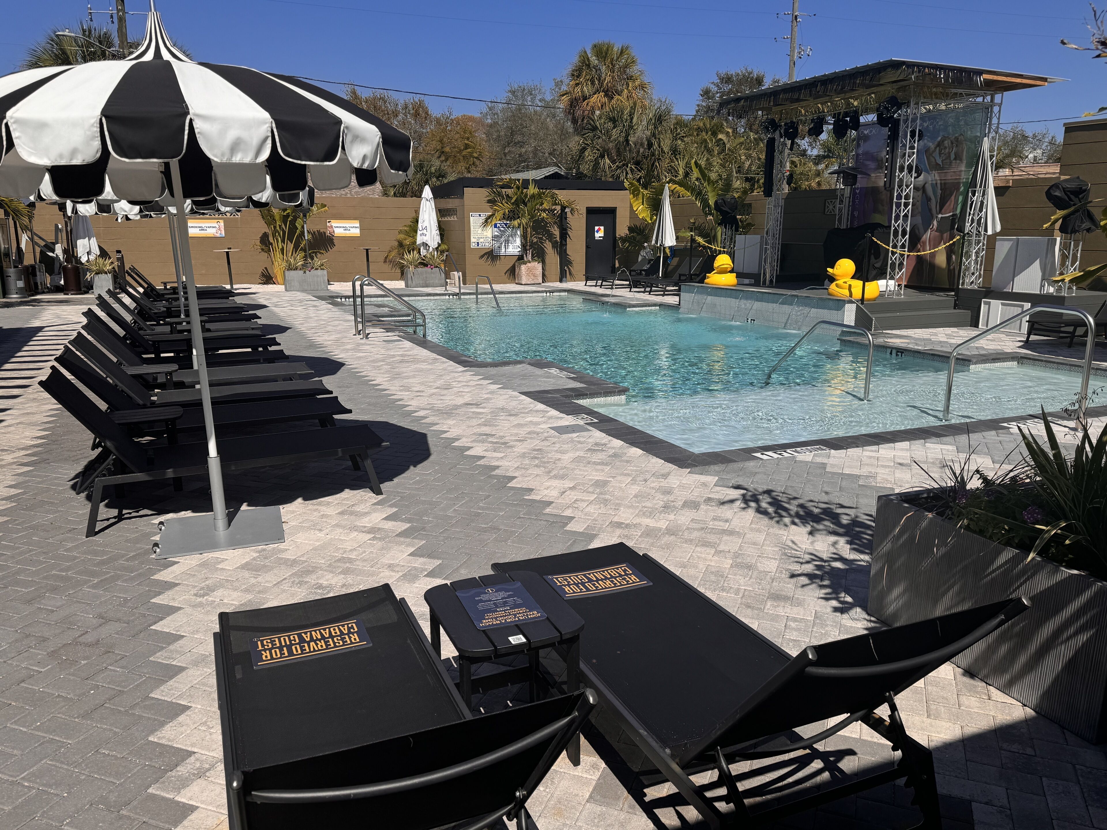 A poolside area with lounge chairs and umbrellas, a swimming pool, and a small stage with speakers.