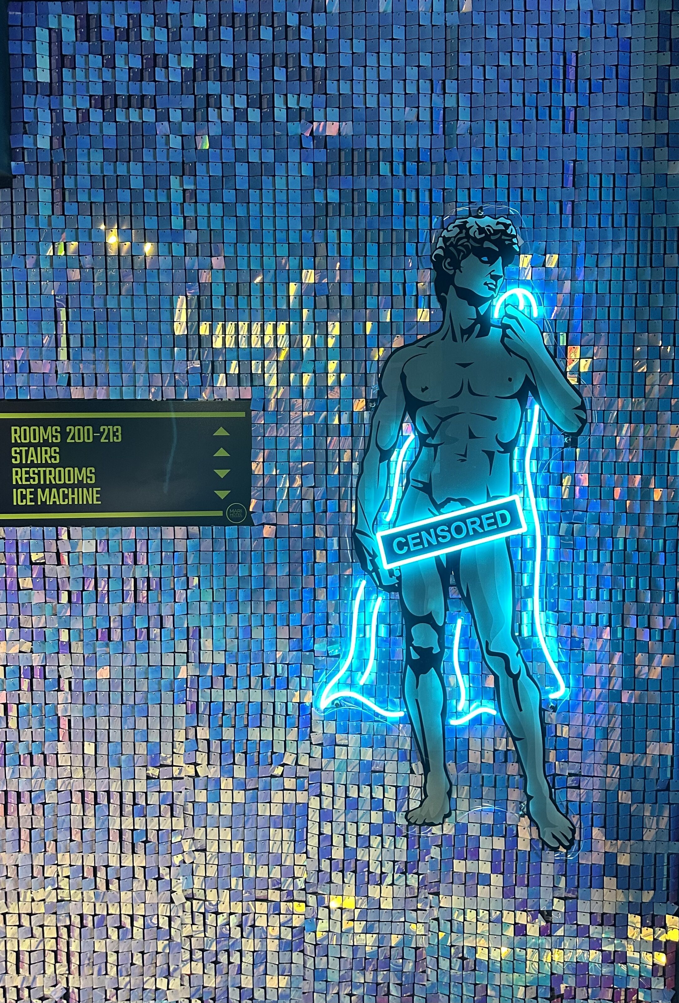 Eine Mosaikwand mit einer Neonreklame, die Annehmlichkeiten wie Zimmer, Treppen, Toiletten und einen Eisbereiter anzeigt.