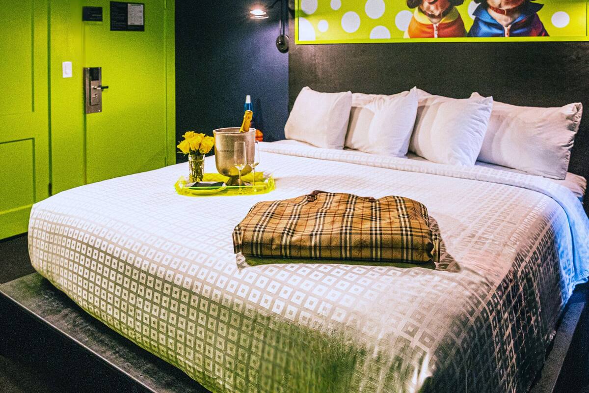 Een hotelkamer met een bed, kussens, een ruitjeskussen, een vaas met bloemen en een fles champagne.