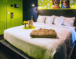 A hotel room with a bed, pillows, a plaid pillow, a vase with flowers, and a bottle of champagne.