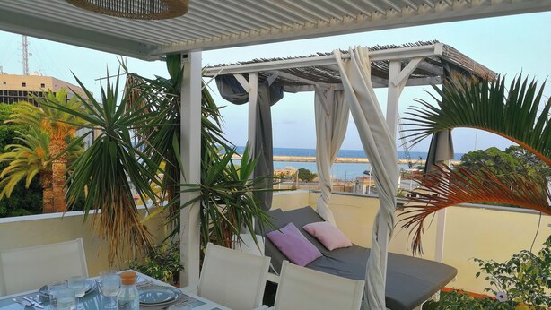 A glass-top dining table set for four with white chairs, a cushioned outdoor seating area, and a view of the sea. - Atlantic Terrace. An oasis in the city. (Santa Cruz de Tenerife)
