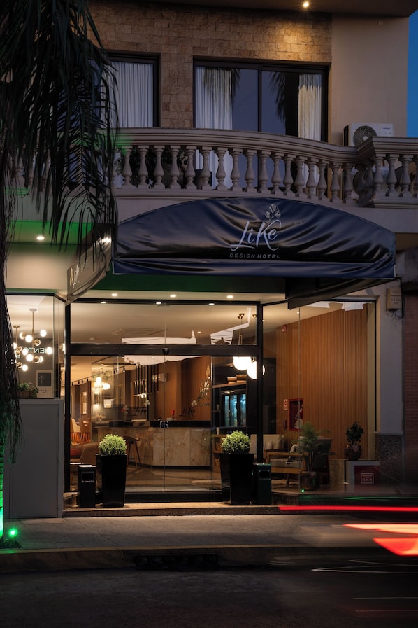 A hotel entrance with a covered outdoor area, a reception desk, and a visible interior with seating and plants.