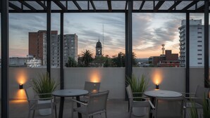 Patio esterno con tavoli e sedie rotondi, piante in vaso e vista sui palazzi e su una torre dell'orologio al crepuscolo.