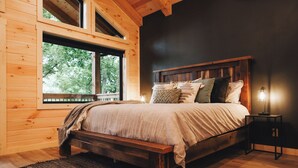 Un dormitorio de cabaña de madera con una cama grande, una mesita de noche y una ventana con vistas a árboles.