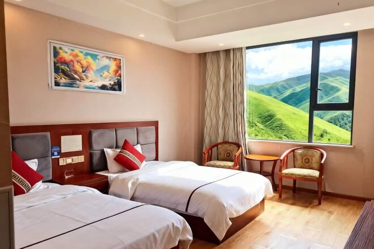A hotel room with two beds, a painting, a view of green hills, a chair, and a small table.