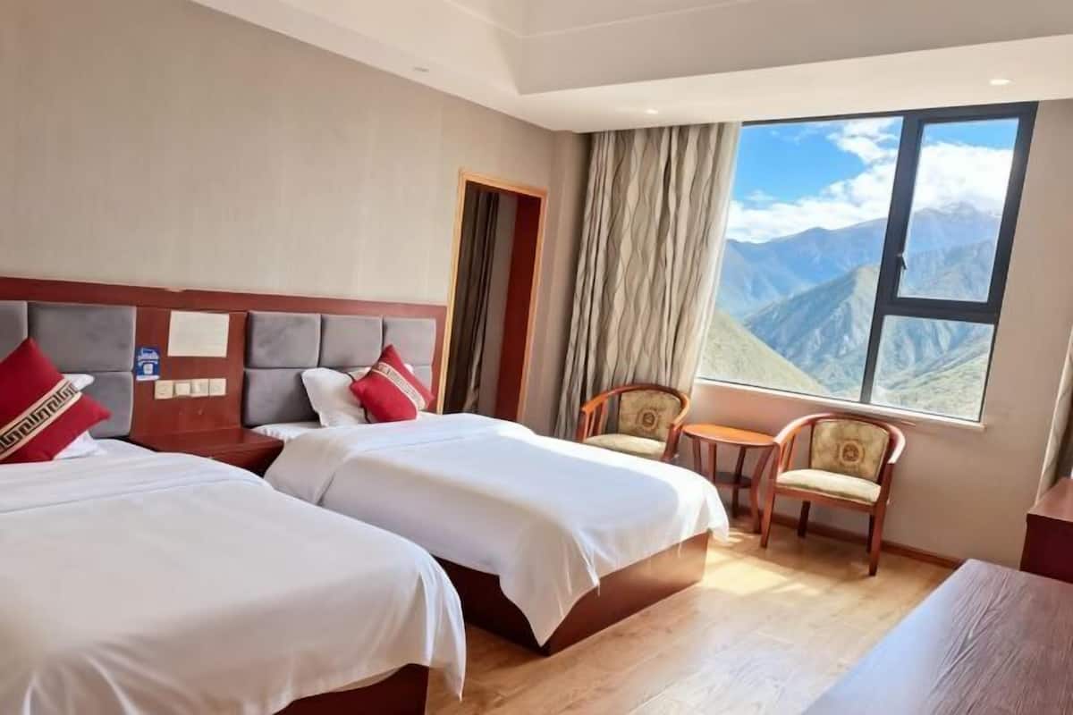 A hotel room with two beds, a small table, a chair, and a large window offering a mountain view.