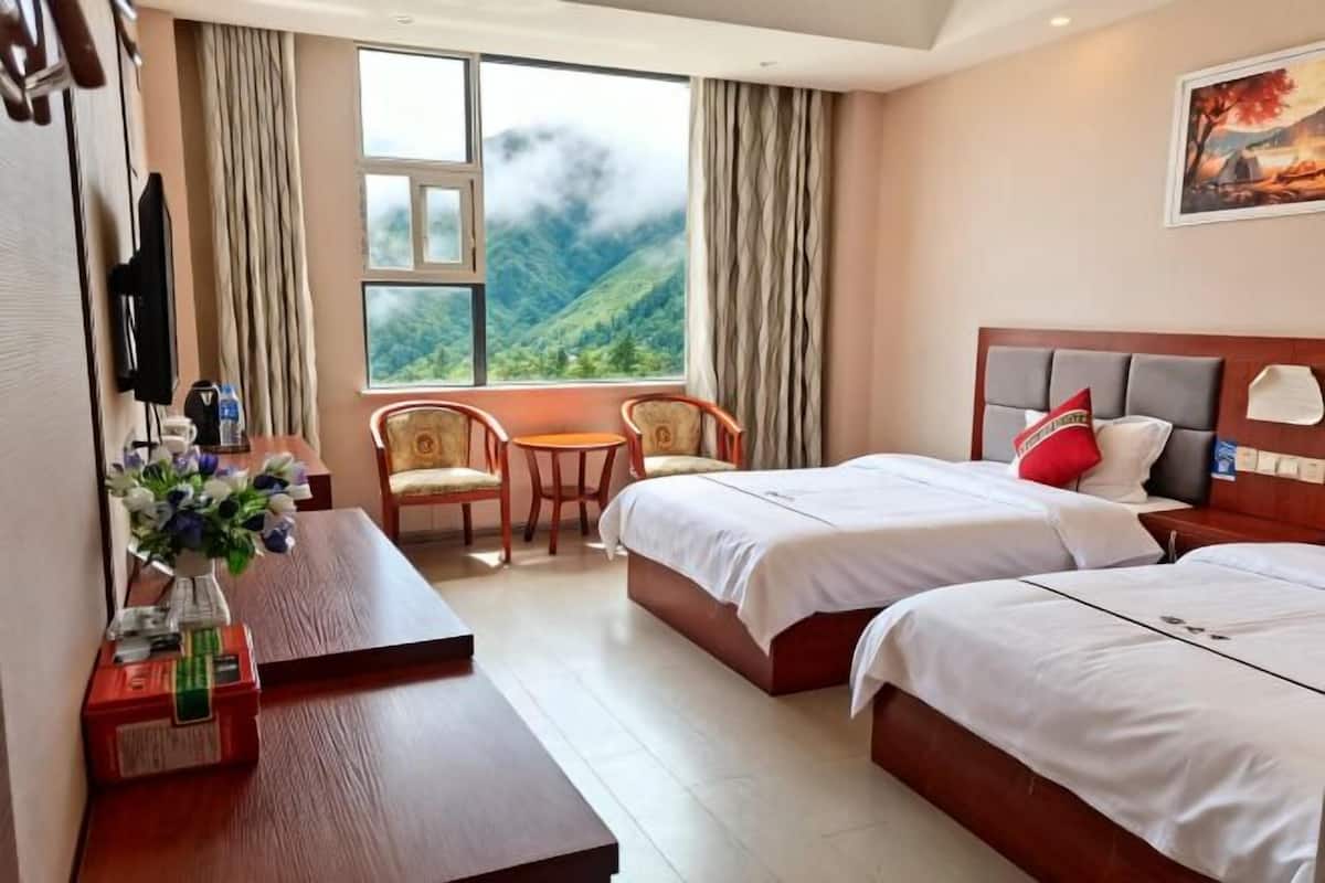 A hotel room with two beds, a desk, a chair, a TV, and a window with a mountain view.