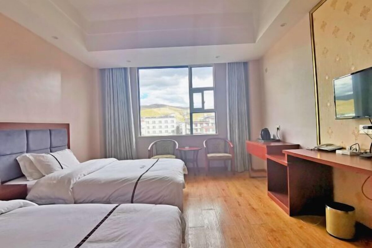 A hotel room with two beds, a desk, a chair, a TV, and a large window with a view of buildings.