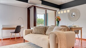 Un salon moderne avec un canapé, un bureau et une chaise. Une grande fenêtre offre une vue sur la verdure à l’extérieur.