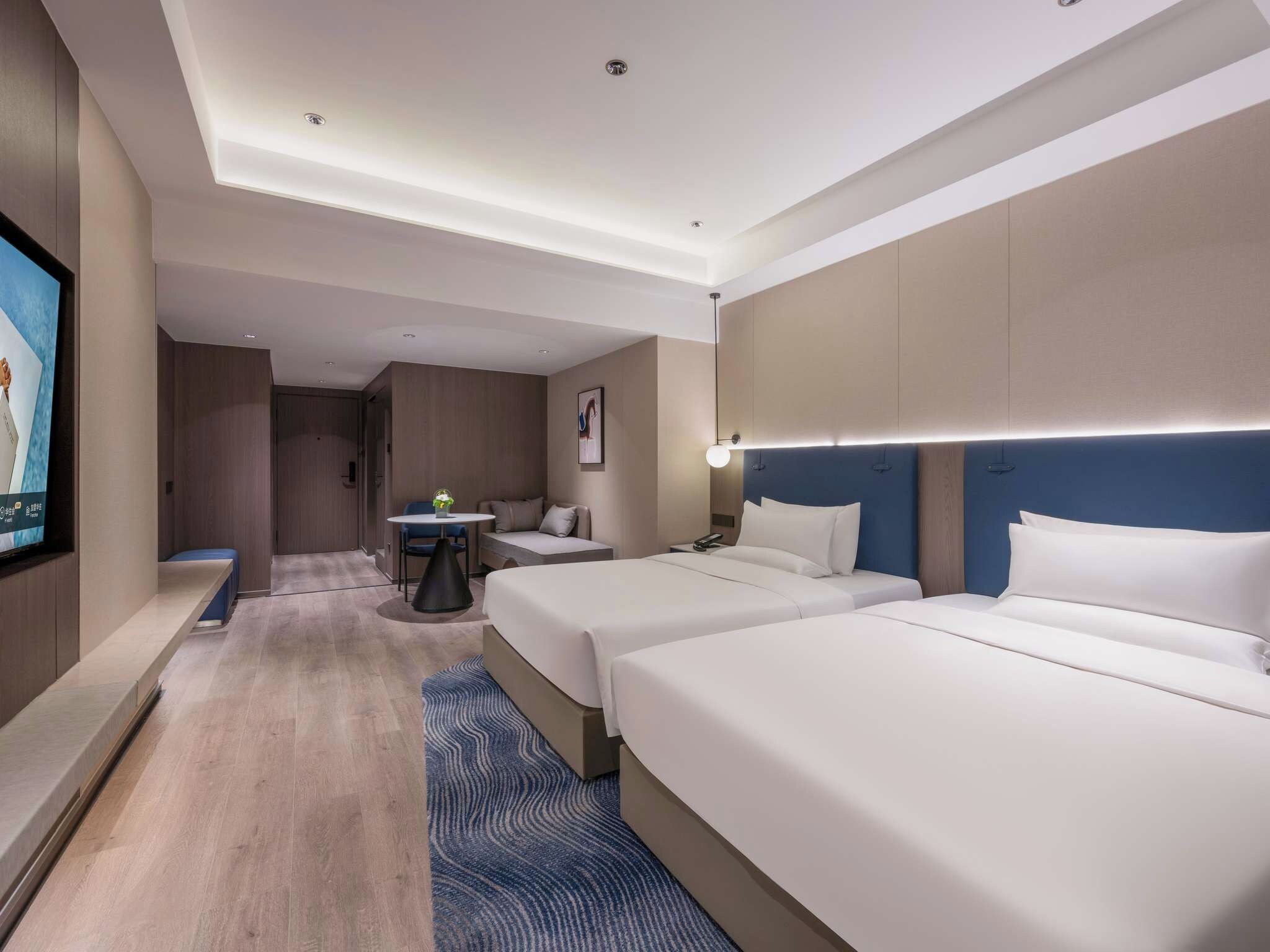 Habitación de hotel con dos camas, un escritorio, una silla, una mesita y un televisor montado en la pared.