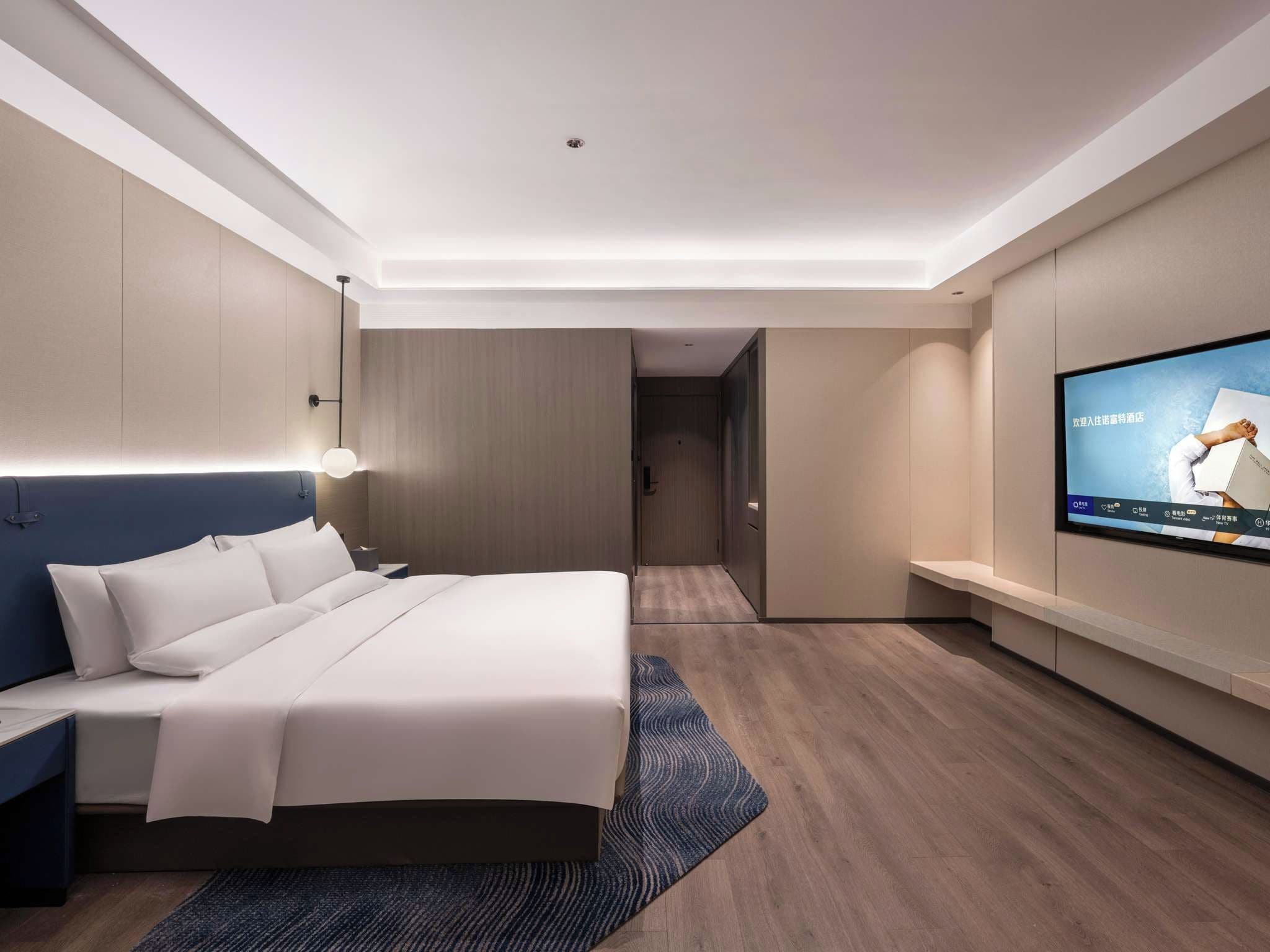 Habitación de hotel moderna con cama, mesita de noche, televisor y vista al pasillo.