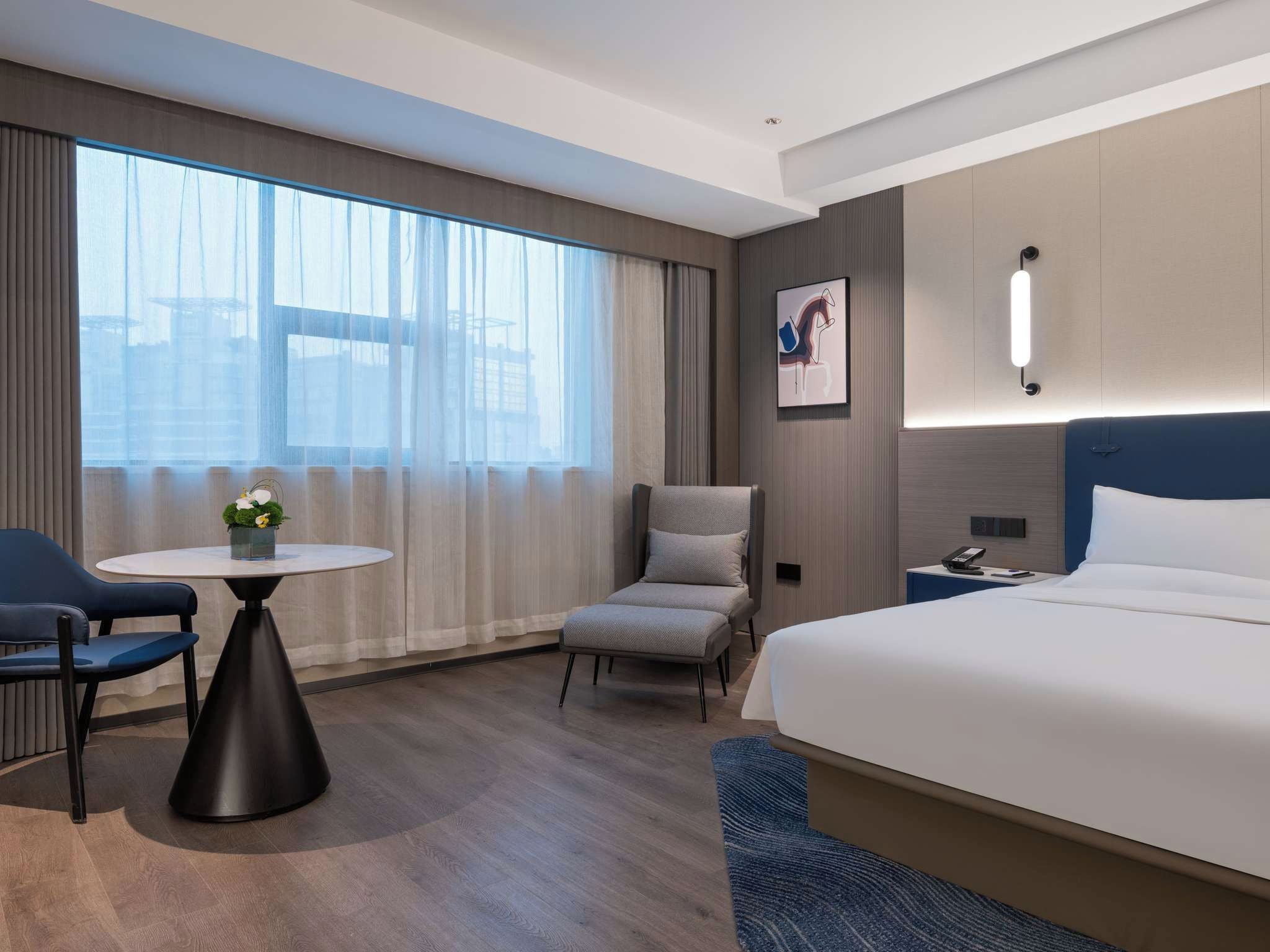 A hotel room with a bed, a chair, a small table with a vase, and a large window with curtains.